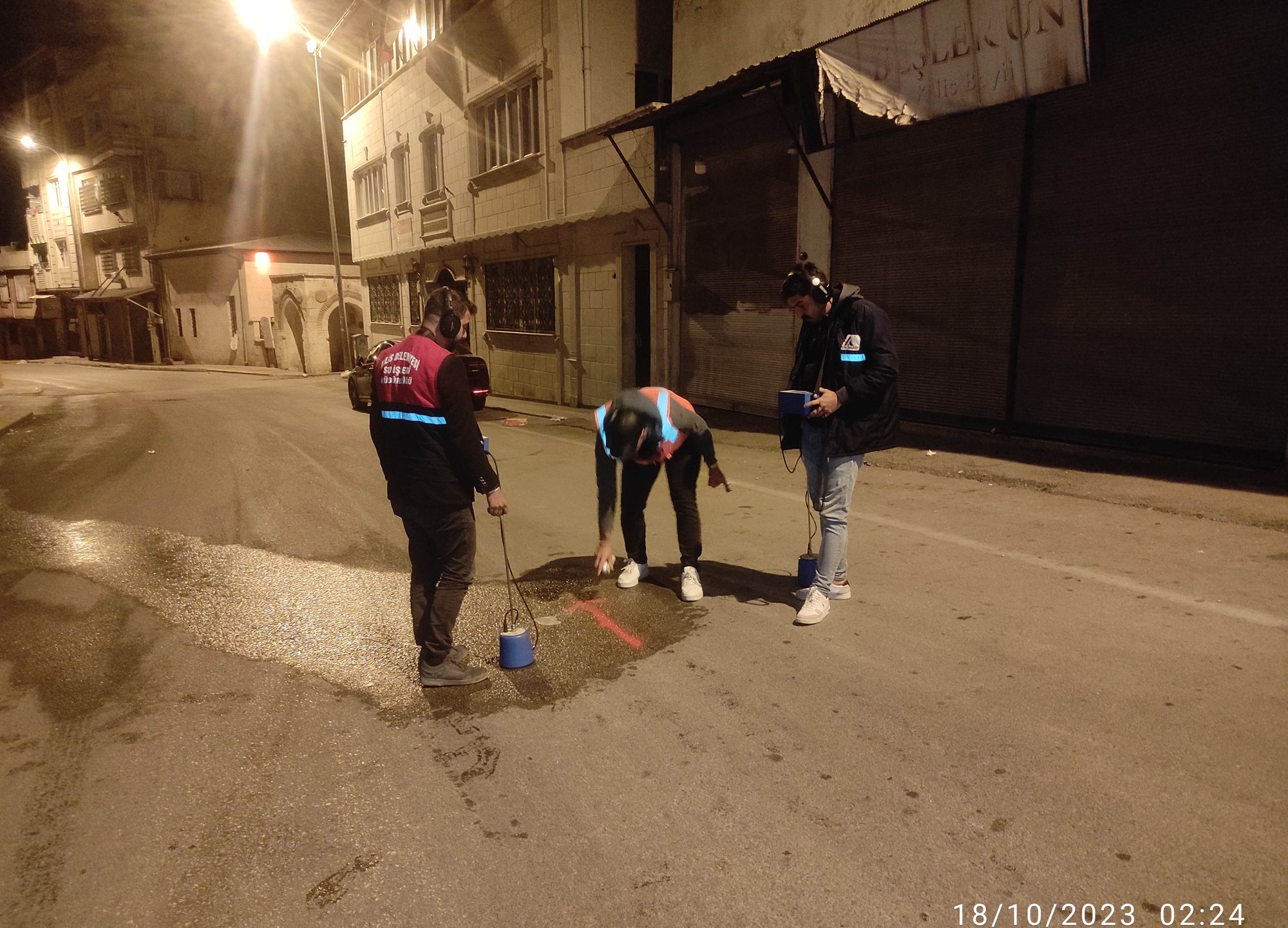 Gece saatlerinde bir cadde üzerinde üç kişilik LeakExpert ekibi kulaklık ve yer mikrofonlarıyla akustik dinleme yaparken, zemindeki ıslak lekeye işaret koyuyor